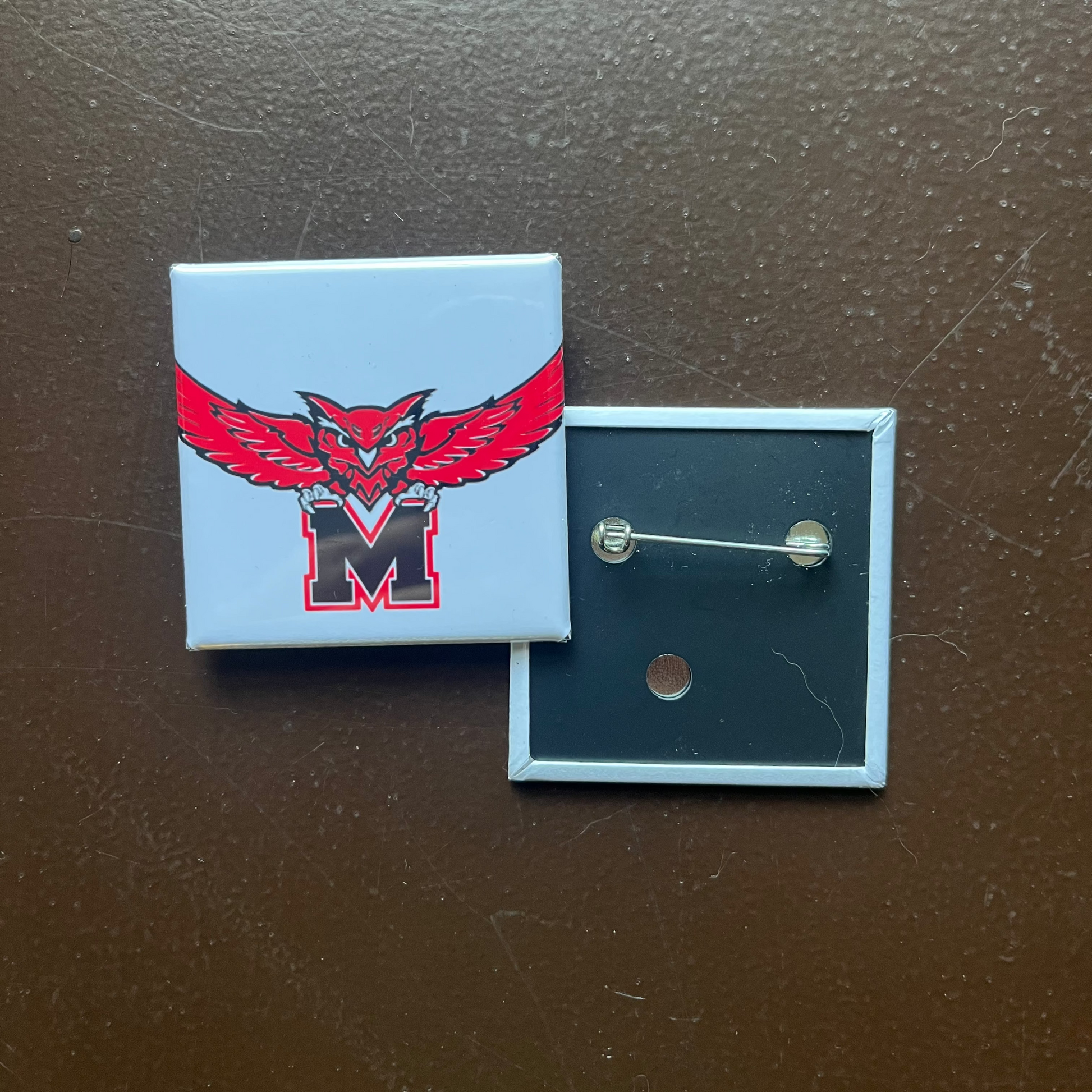 Pin with a red and black logo on a blue backing, on a dark brown surface.